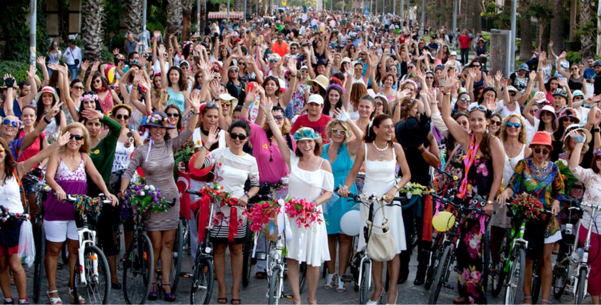 A dense crowd of hundreds of smiling and waving women with their decorated bicycles at the start of the ride. They wear colourful, sleeveless summer dresses, broad-brimmed hats, and attached to their bike handlebars are large bouquets of flowers. 
